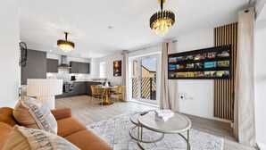 A modern living room with a sofa, a coffee table, and a dining area with a table and chairs. There is a kitchen in the background, and a TV mounted on the wall displaying various channels. - Brandnew Luxurious 2bedApartment (Ebbsfleet Valley)
