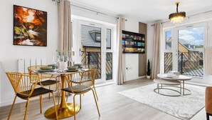 A modern dining area with a round table, gold chairs, and a large window with a view of buildings. - Brandnew Luxurious 2bedApartment (Ebbsfleet Valley)