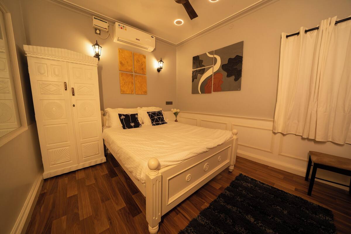 Une chambre avec un lit blanc, un plancher en bois, un tapis noir, une armoire blanche et un tableau accroché au mur.