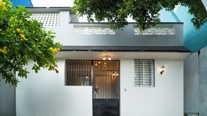 A white building with a black gate, a small window with bars, and a tree with yellow flowers. - Eden Garden By Jolly Homes (Madurai)