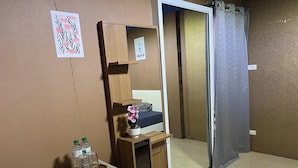 A compact room with a bed, a small wooden shelf with a potted orchid, a mirror, and a refrigerator.