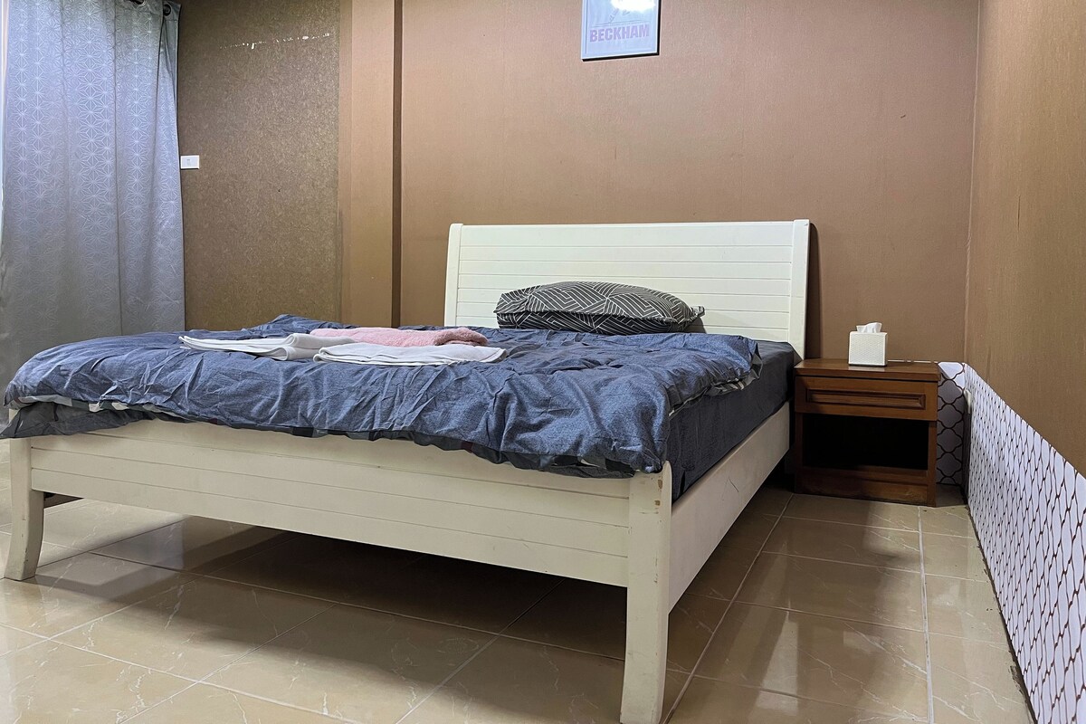A neatly made bed with a white headboard, a bedside table with a tissue box, and a framed picture on the wall.