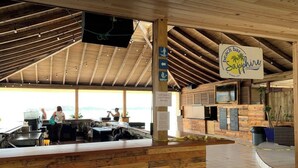 Een strandbar met houten constructie, een bar met een menubord en een bordje dat de herentoiletten aangeeft.