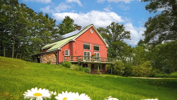 A red barn with a stone foundation, a green roof, and a wooden deck. - Charming Barn Home Near Jiminy Peak (Hancock)