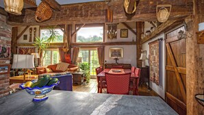 A rustic dining area with a wooden table and chairs, a large window with curtains, and a view of greenery outside. - Charming Barn Home Near Jiminy Peak (Hancock)
