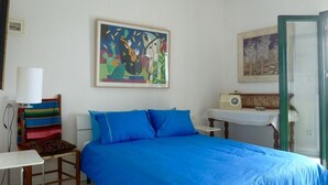 A bedroom with a large bed, a chair with a colorful blanket, a desk, a radio, and two paintings on the wall.