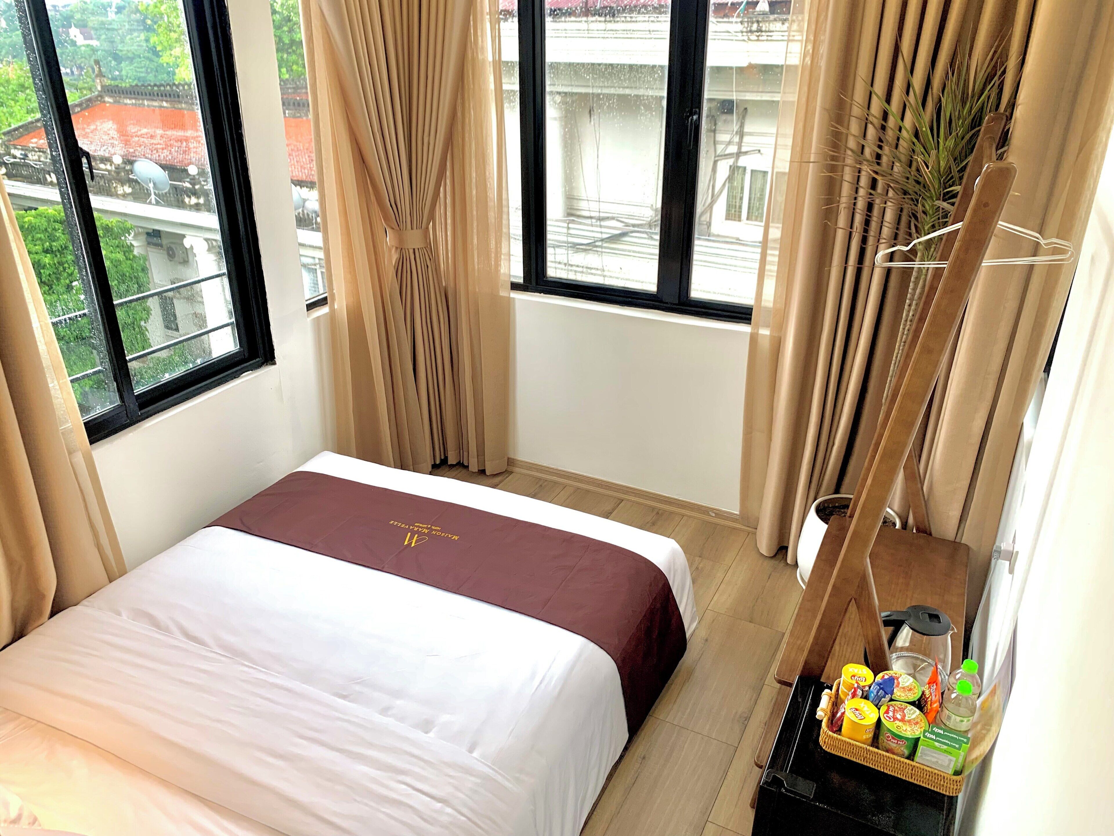 A hotel room with a bed, a window with curtains, a small table with toiletries, and a view of buildings outside.
