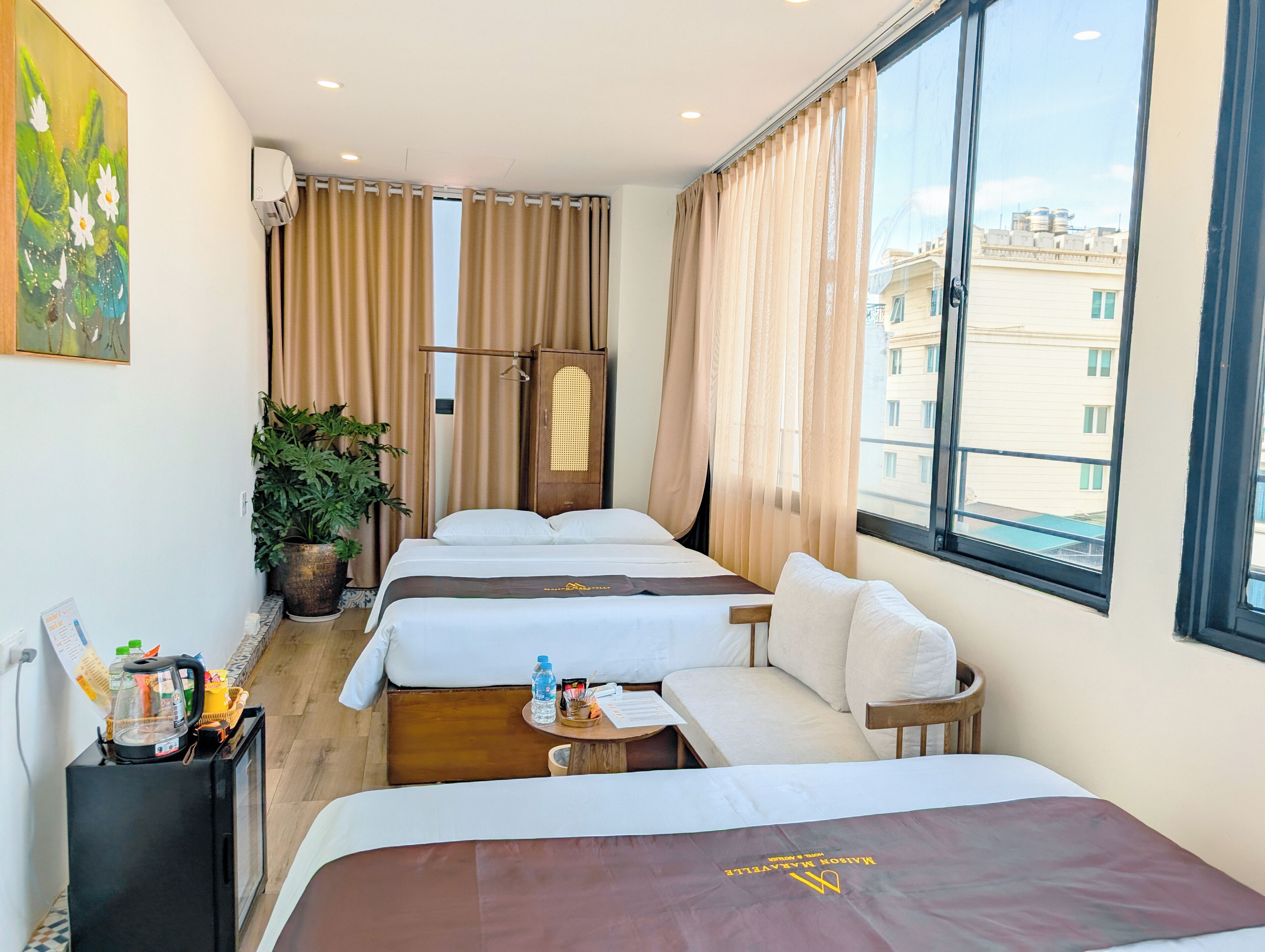 A hotel room with two beds, a sofa, a small table, a potted plant, and a window with a view of buildings.