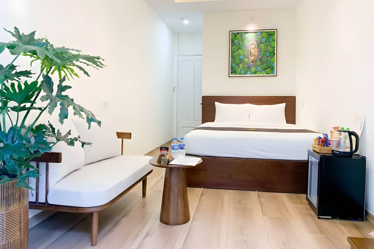 A hotel room with a bed, a chair, a small table, a potted plant, a painting, and a coffee machine.