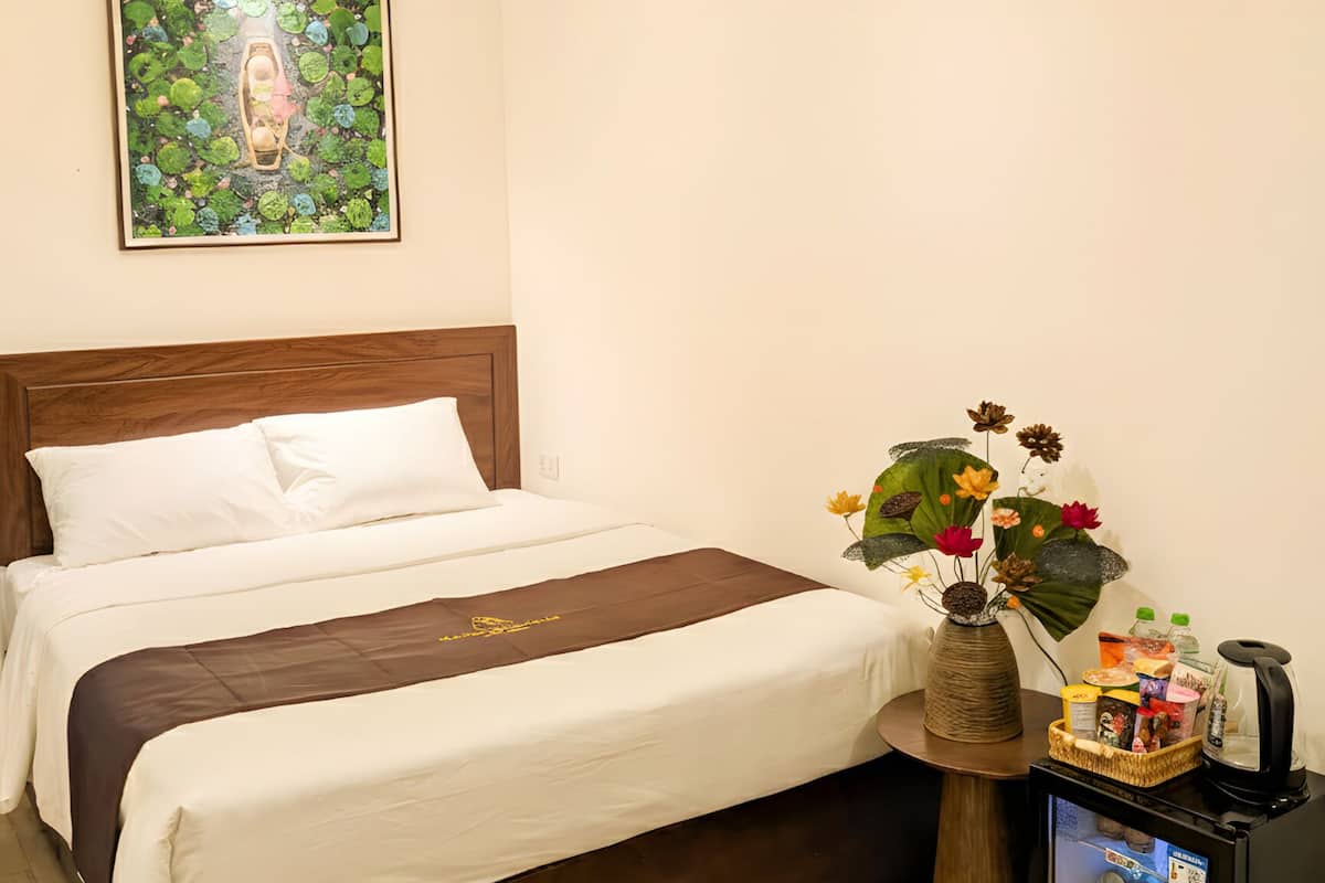 A neatly arranged hotel room with a bed, a small table with a vase of flowers, a refrigerator, and a painting on the wall.