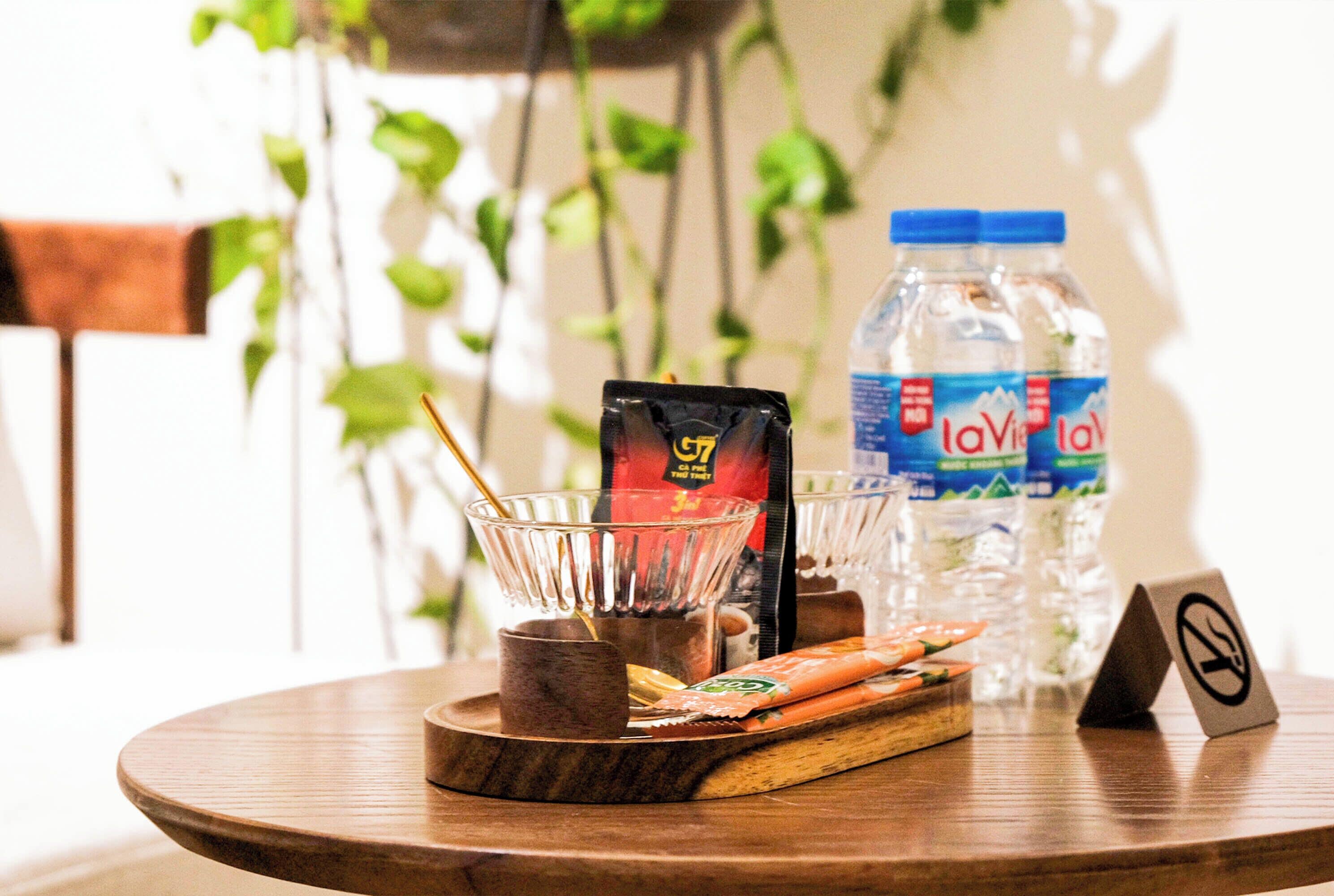 A wooden table with a glass bowl containing a packet of snacks, a small cup, and a wooden tray with snacks. There are two bottles of water and a no-smoking sign on the table.