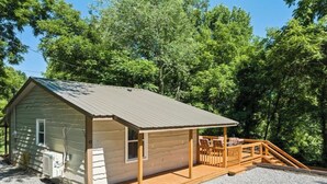 A small cabin with a wooden deck and a covered porch, surrounded by trees.