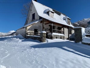 Une maison recouverte de neige, avec des fondations en pierre, un porche en bois et un ciel d’un bleu limpide.