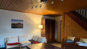 Una stanza accogliente con soffitto in legno, un tavolo grande, due divani con cuscini, una lampada e una porta.