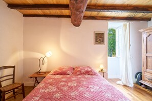 Un dormitorio con techo de madera, una cama con colcha floral, una silla, una mesita de noche con lámpara y un armario.