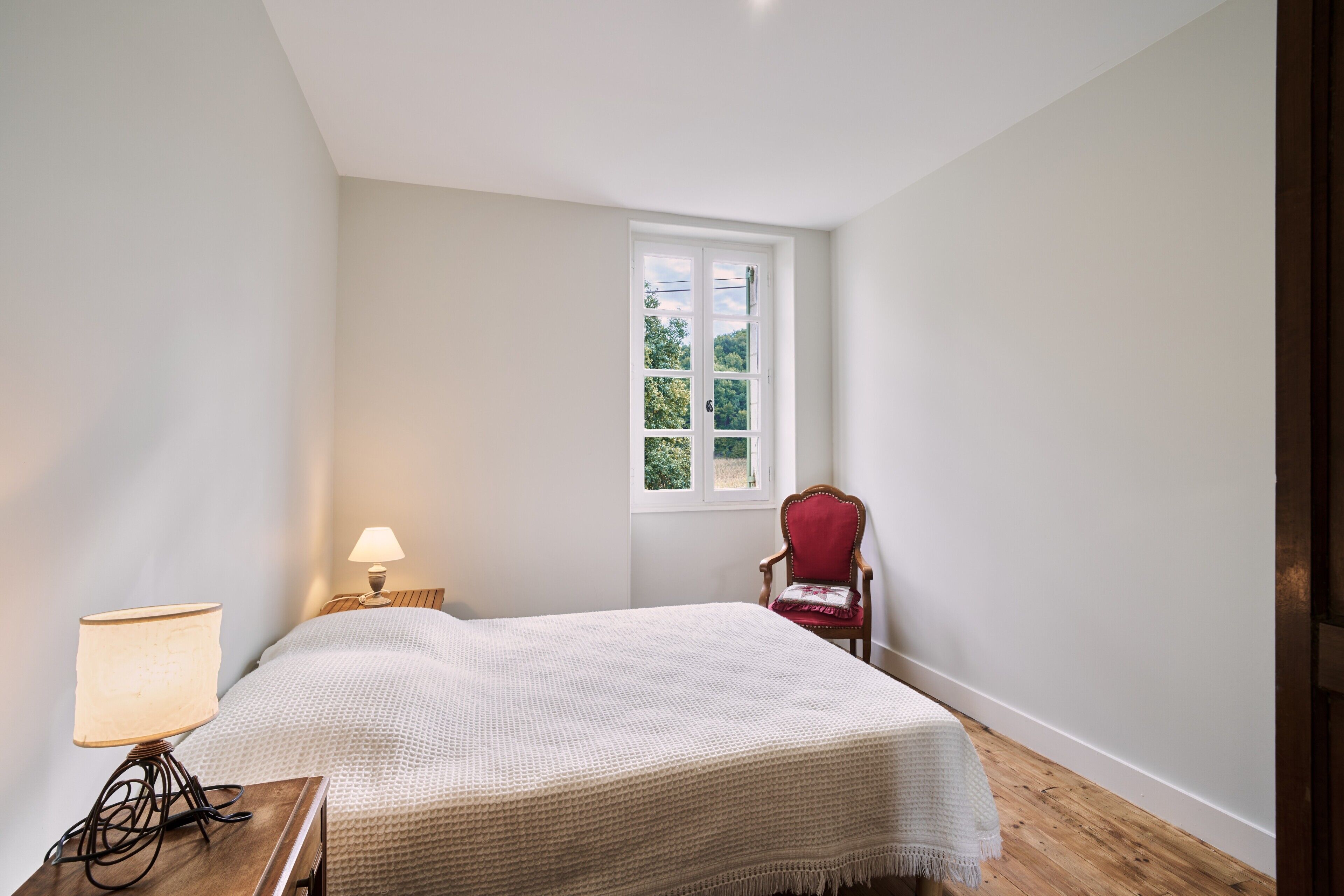 A bedroom with a bed, a nightstand with a lamp, a chair, and a window.