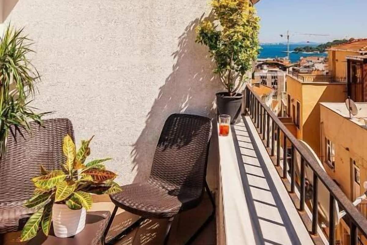 A balcony with wicker furniture, a potted plant, and a clear view of the sea and buildings.
