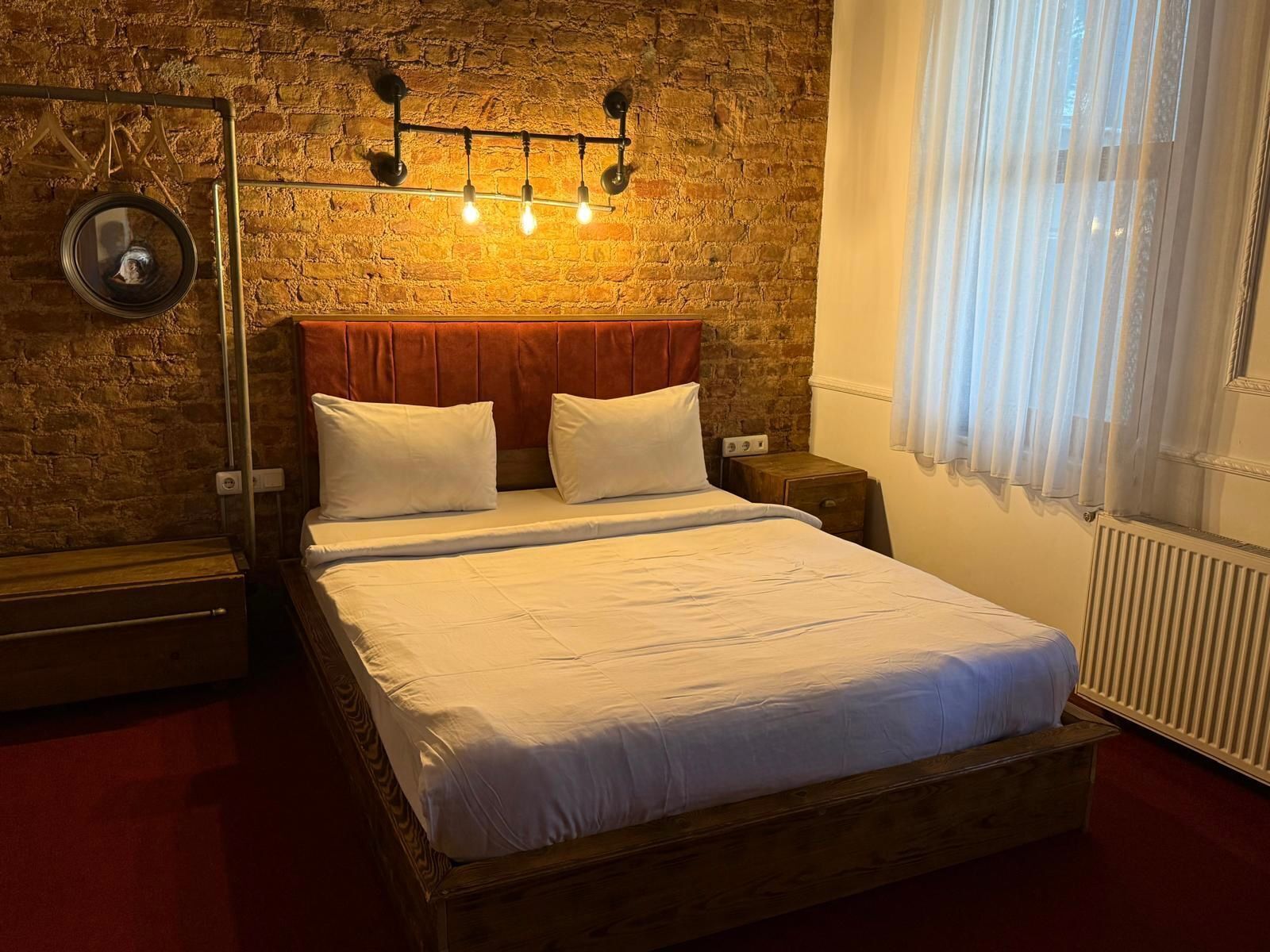 Un dormitorio con pared de ladrillo, estructura de cama de madera, ropa de cama blanca, una mesita de noche y un aplique de pared.