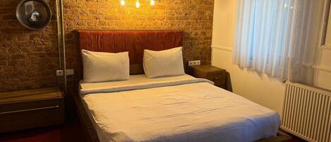 Un dormitorio con pared de ladrillo, estructura de cama de madera, ropa de cama blanca, una mesita de noche y un aplique de pared.