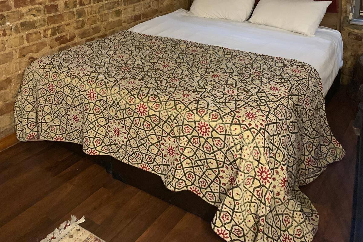 A room with a brick wall, a bed with a patterned bedspread, a mounted TV, and a window with blinds.