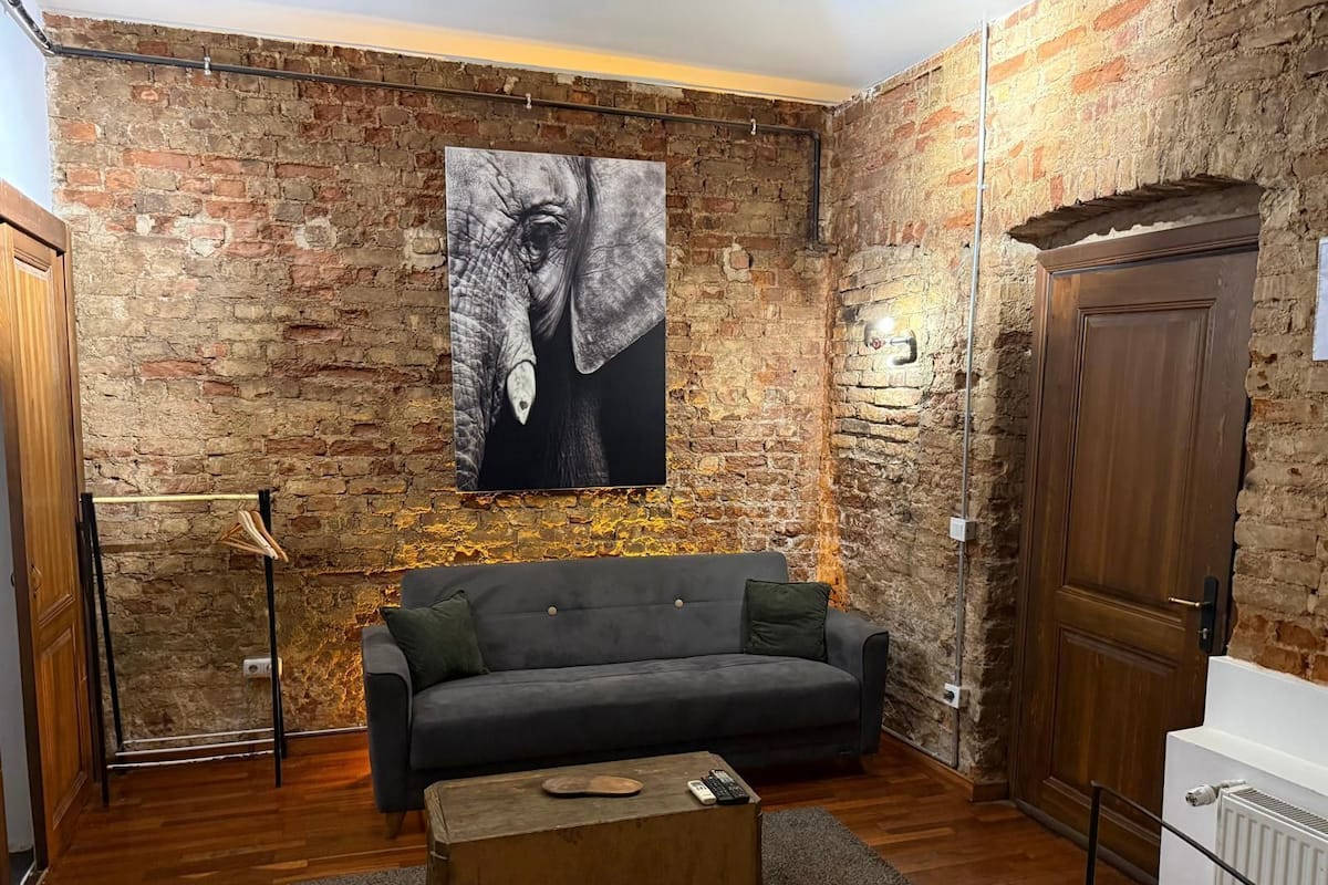 A room with a brick wall, a grey sofa, a wooden coffee table, and a large elephant painting.