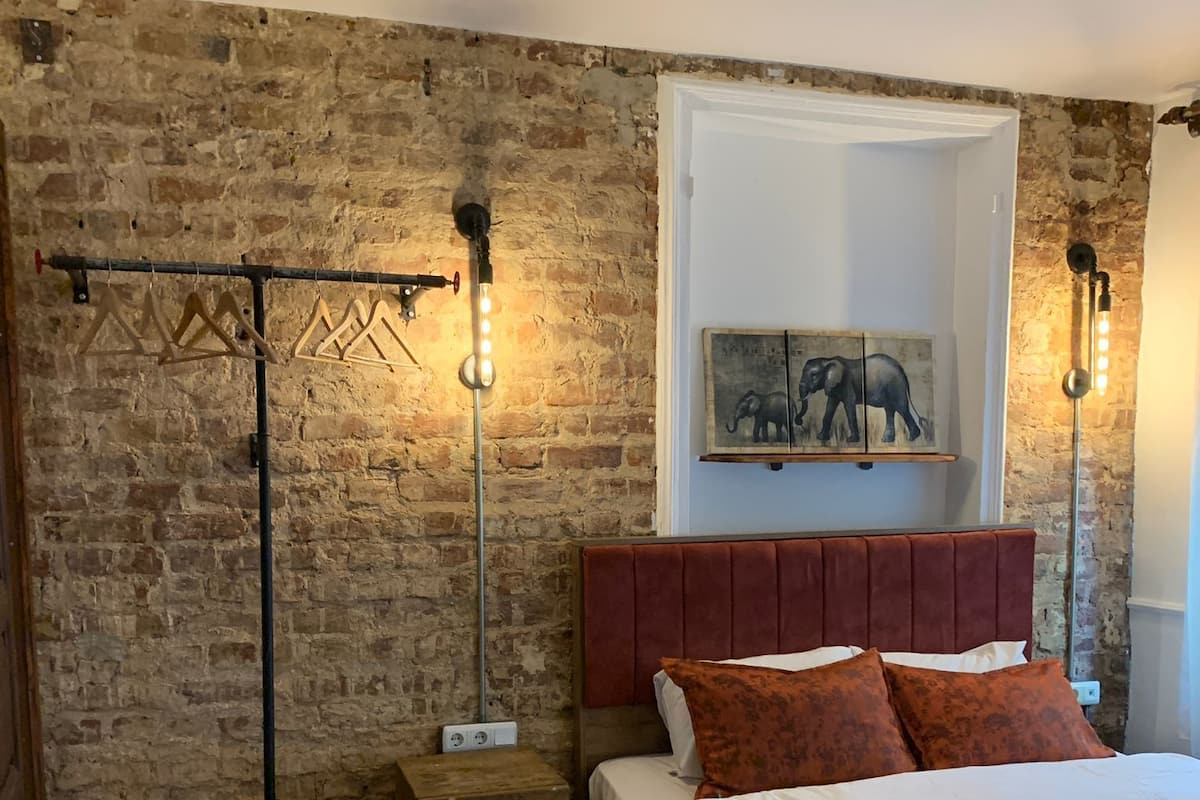 A bedroom with a brick wall, a wooden floor, a bed with a red headboard, and two wall-mounted light fixtures.