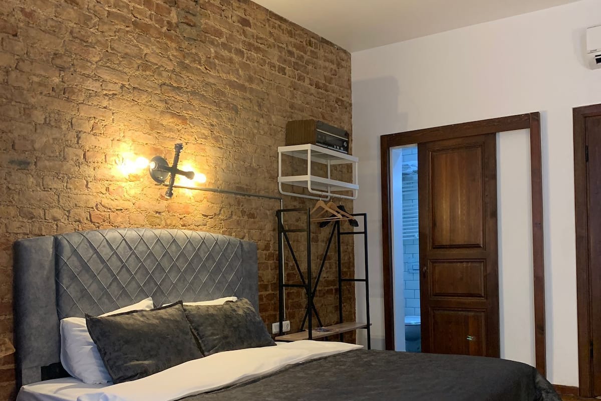 A bedroom with a brick wall, a bed with a grey headboard, a hanging light fixture, and a wooden door.