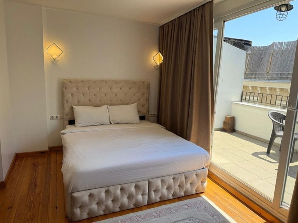 A bedroom with a tufted headboard, a large bed, a sliding glass door to a balcony, and two wall-mounted light fixtures.