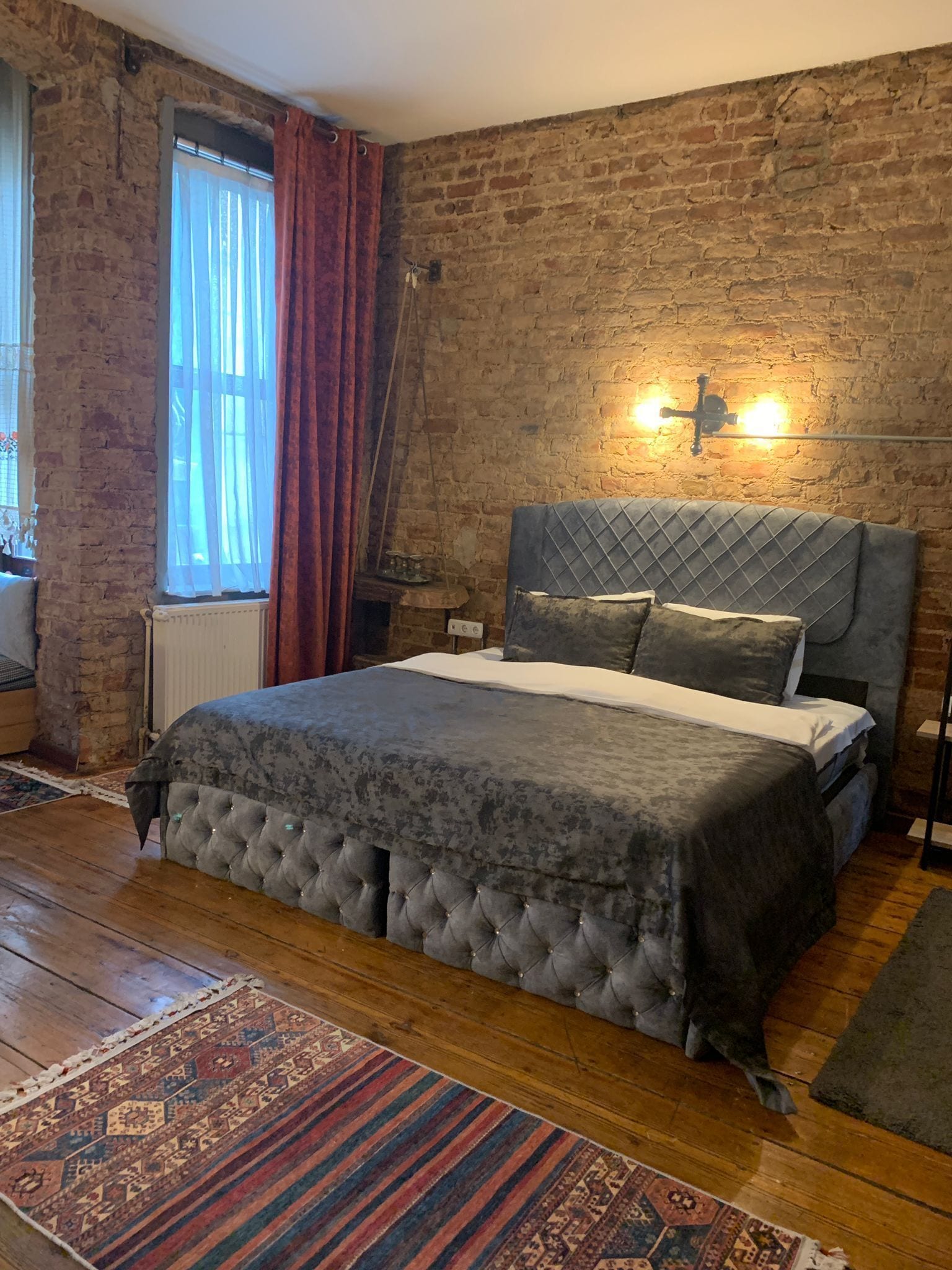 Un dormitorio con pared de ladrillo, una cama con cabecera acolchada, una mesita de noche, una lámpara y una ventana con cortinas.