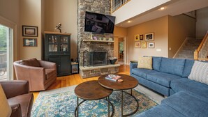 A spacious living room with a stone fireplace, a large flat-screen TV, and a blue sofa. - Lakefront Luxury Chalet w/ Hot Tub & Dock (McHenry)