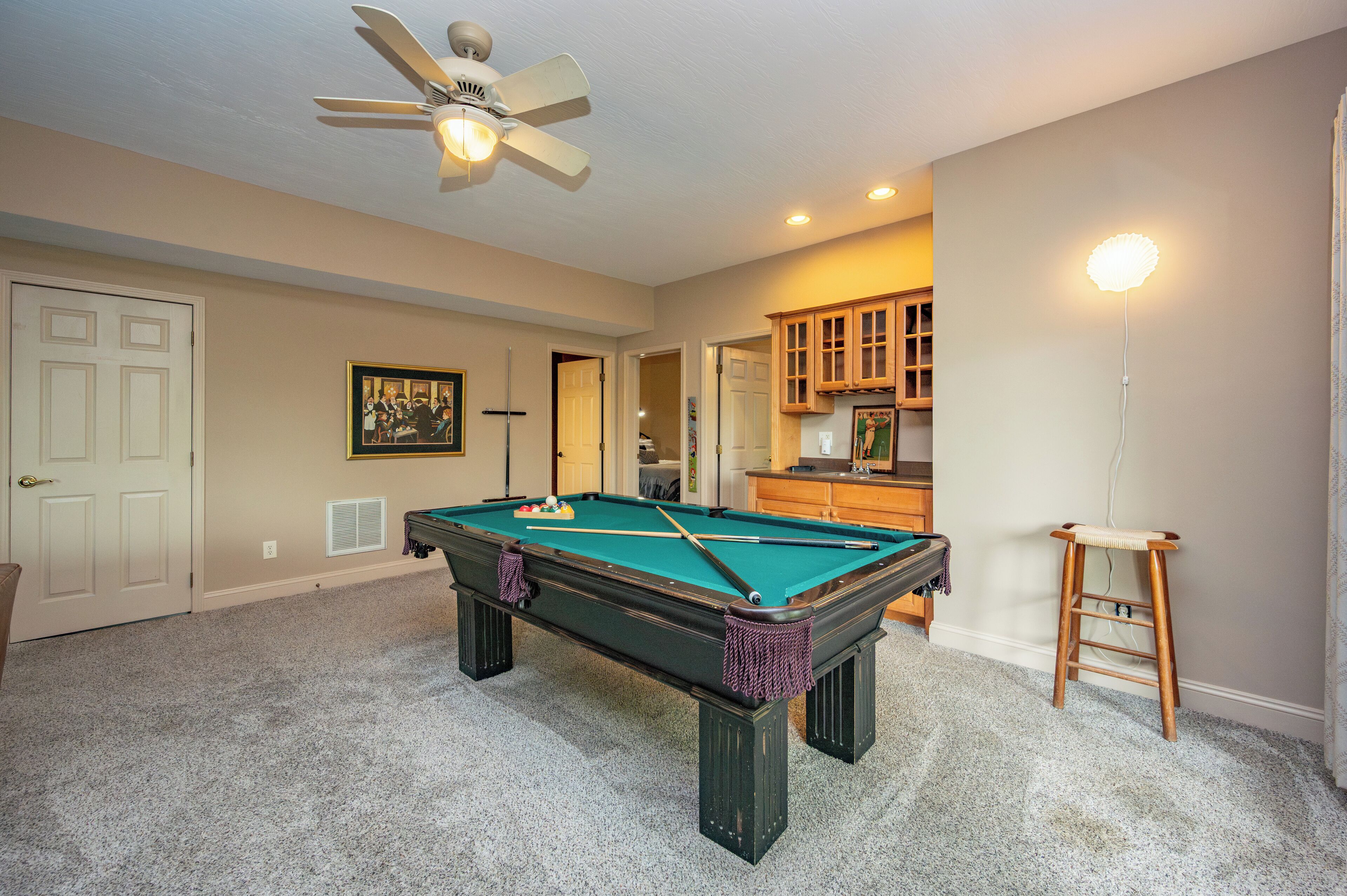A pool table in a room with beige walls, a ceiling fan, and a small stool.