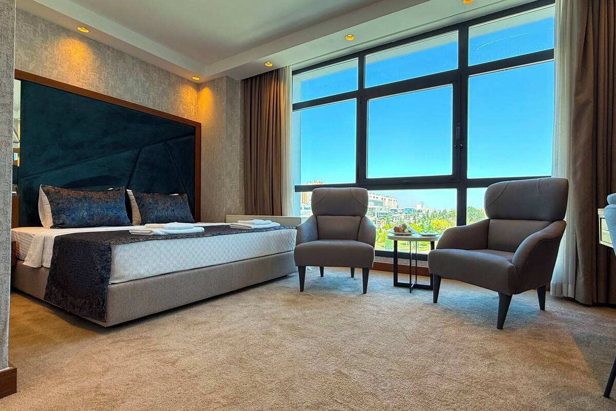 Habitación de hotel con una cama grande, dos sillones, una mesita y un amplio ventanal con vistas a la ciudad.