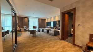 A spacious hotel room with a large bed, a sofa, a dining area with chairs and a table, and a view of the city through large windows. - ALSANCAK PRIME HOTEL KONYA  (Konya)