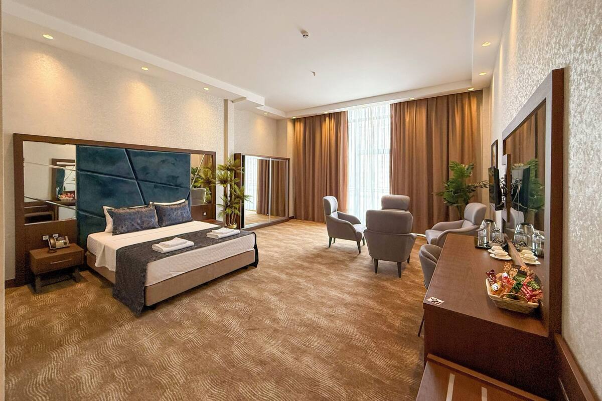 Habitación de hotel moderna con una cama grande, un escritorio con espejo, dos sillones y un ventanal con cortinas.
