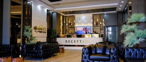 Zona de recepción de hotel con diseño moderno, que incluye un mostrador de recepción, asientos y una pantalla de televisión.