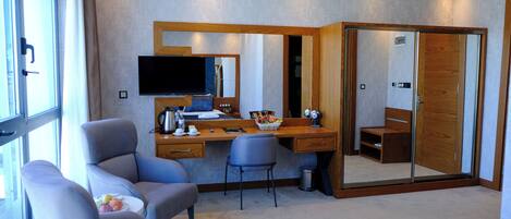 A modern hotel room with a large desk, a comfortable armchair, a television, and a mirror.