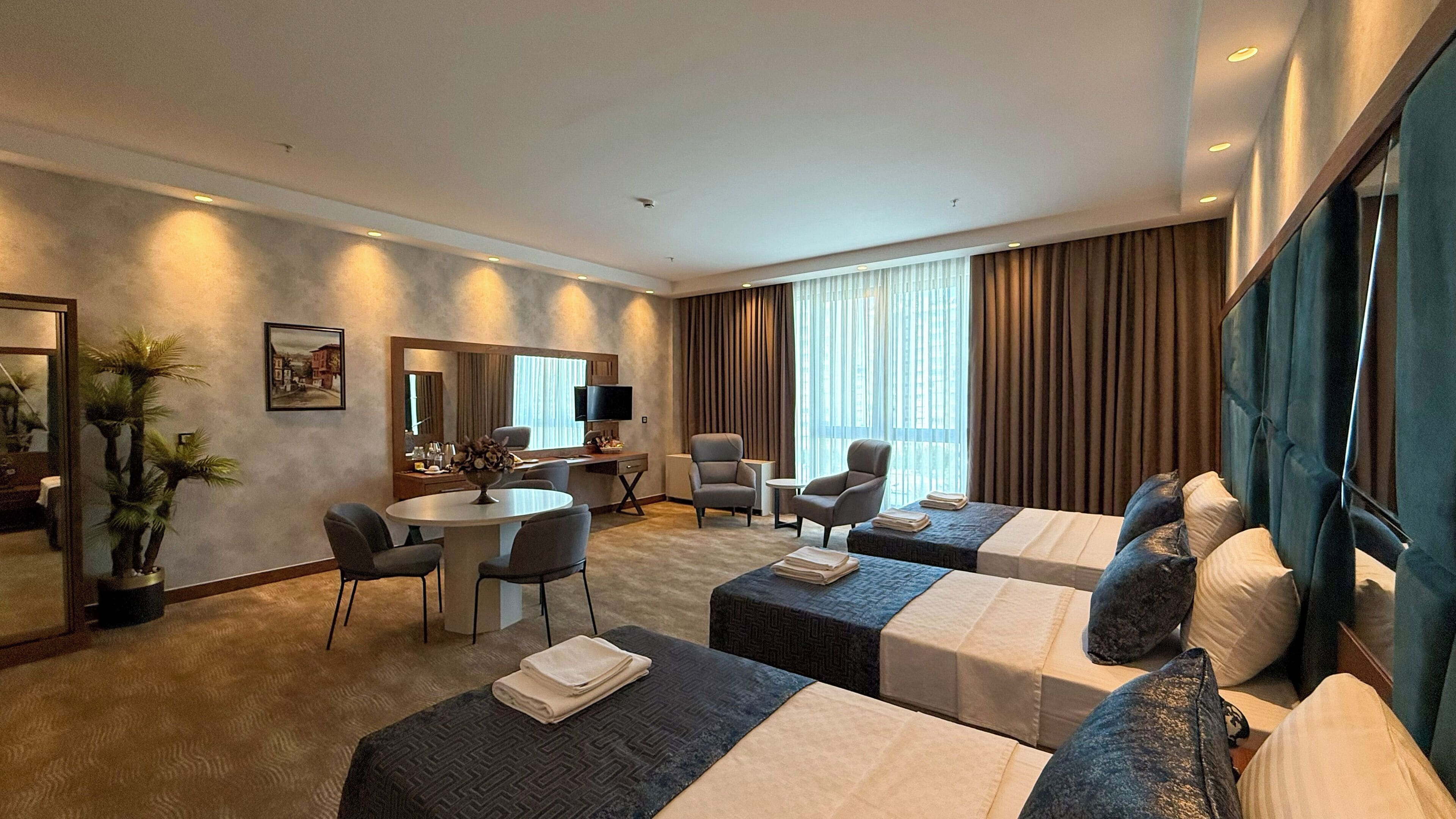 A hotel room with two beds, a desk, chairs, a TV, and a large window with curtains.