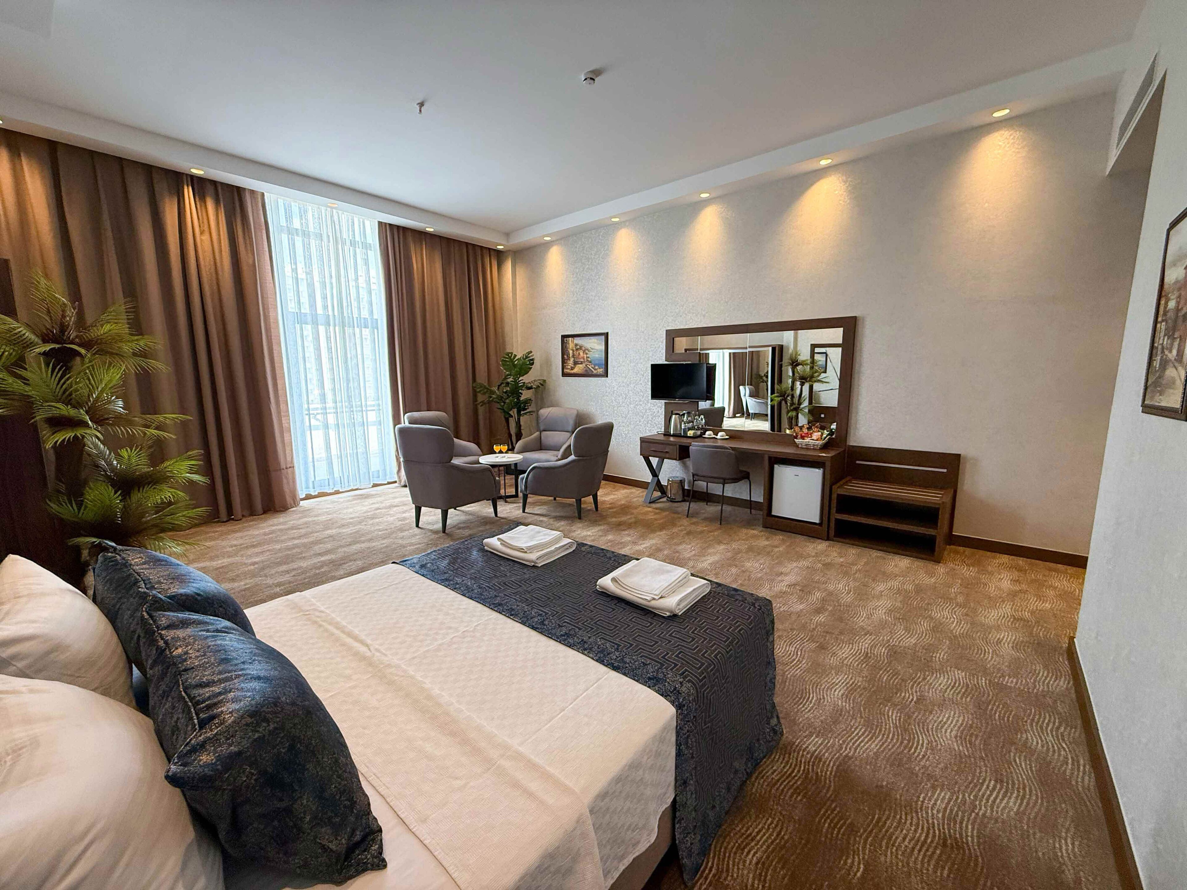 A hotel room with a bed, two armchairs, a desk with a computer, a small refrigerator, and a large window with curtains.