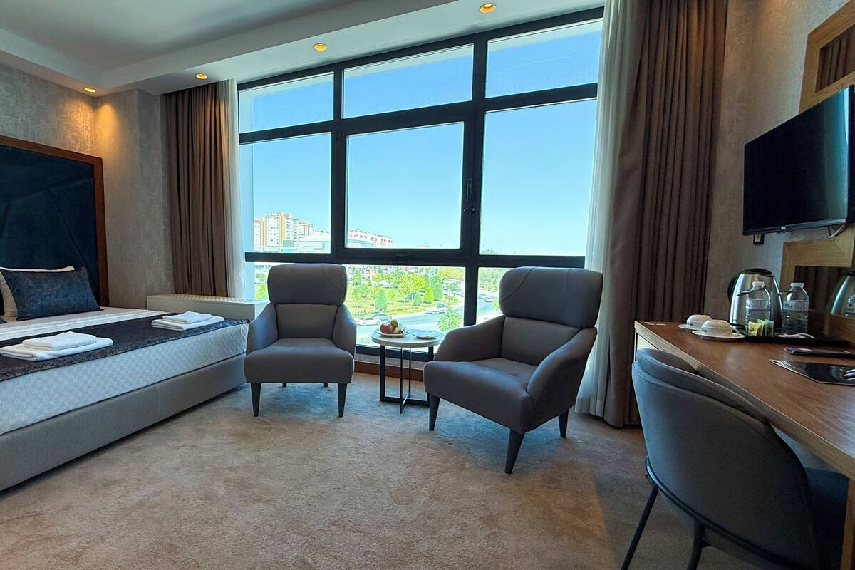 Habitación de hotel con cama, dos sillones, una mesita, un escritorio con televisor y un ventanal con vistas a la ciudad.