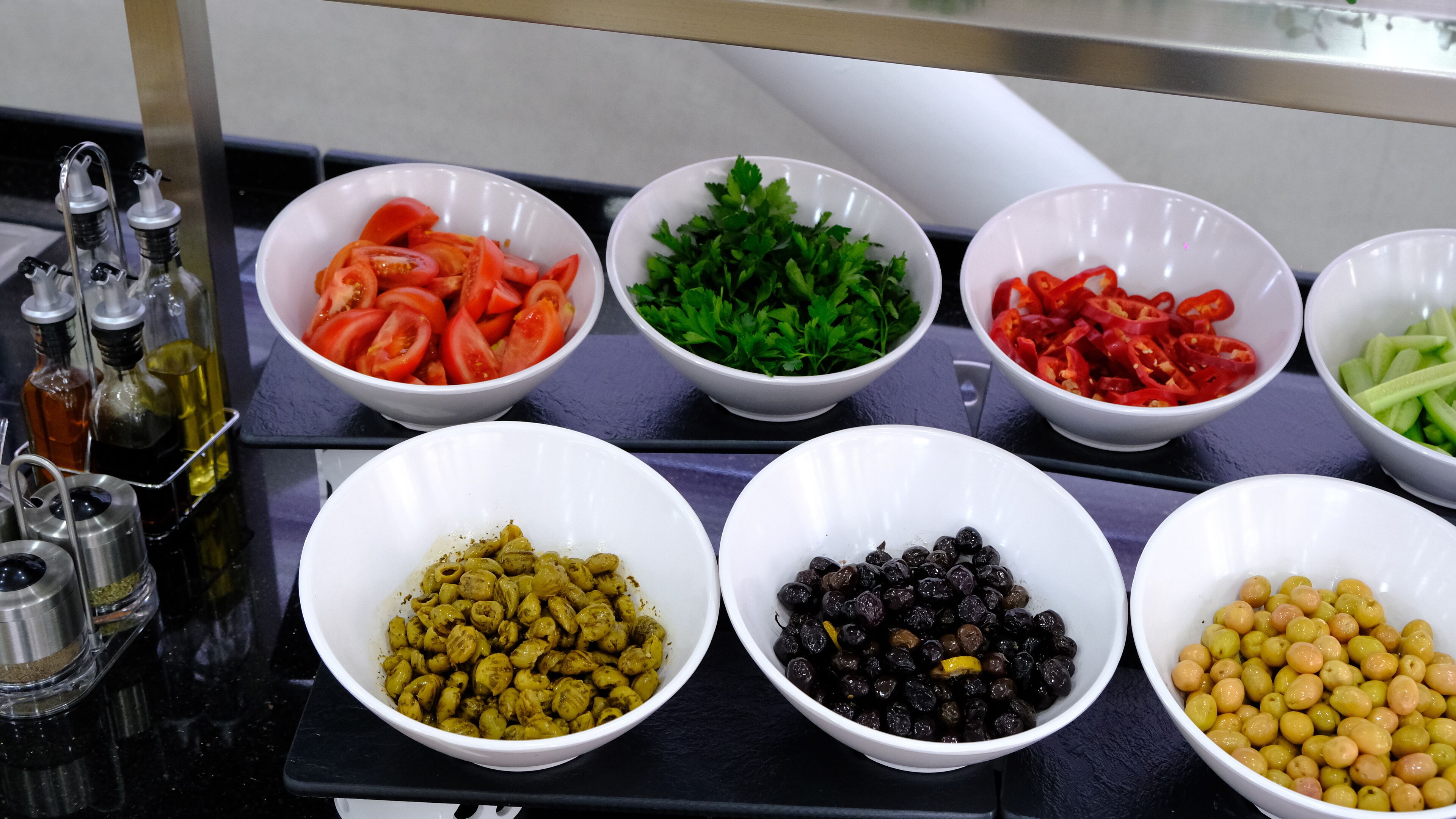 A kitchen counter with various bowls containing ingredients like tomatoes, parsley, peppers, cucumbers, olives, and capers.