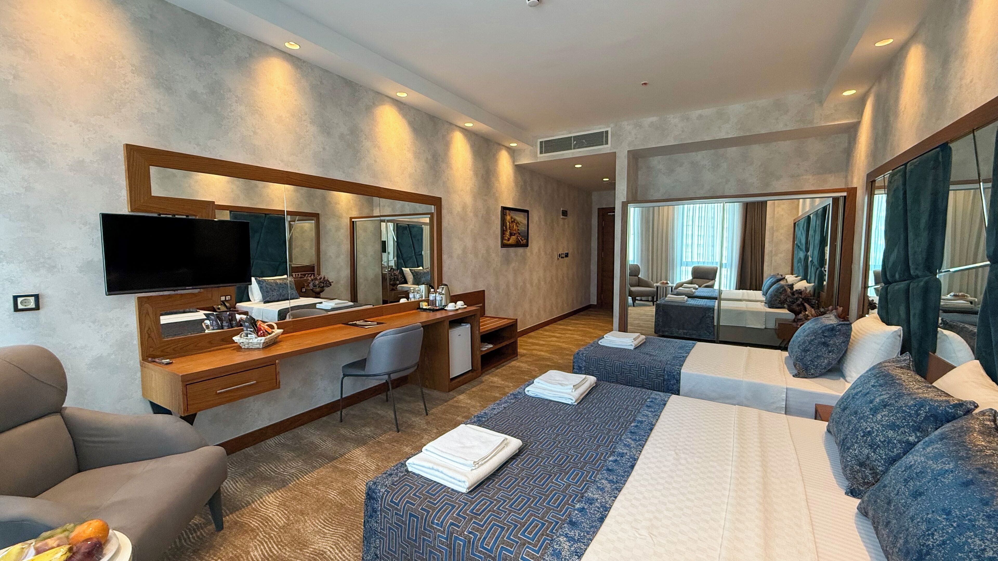 Una habitación de hotel moderna con una cama grande, un escritorio con computadora, televisor y un área de descanso con sofá y sillones.