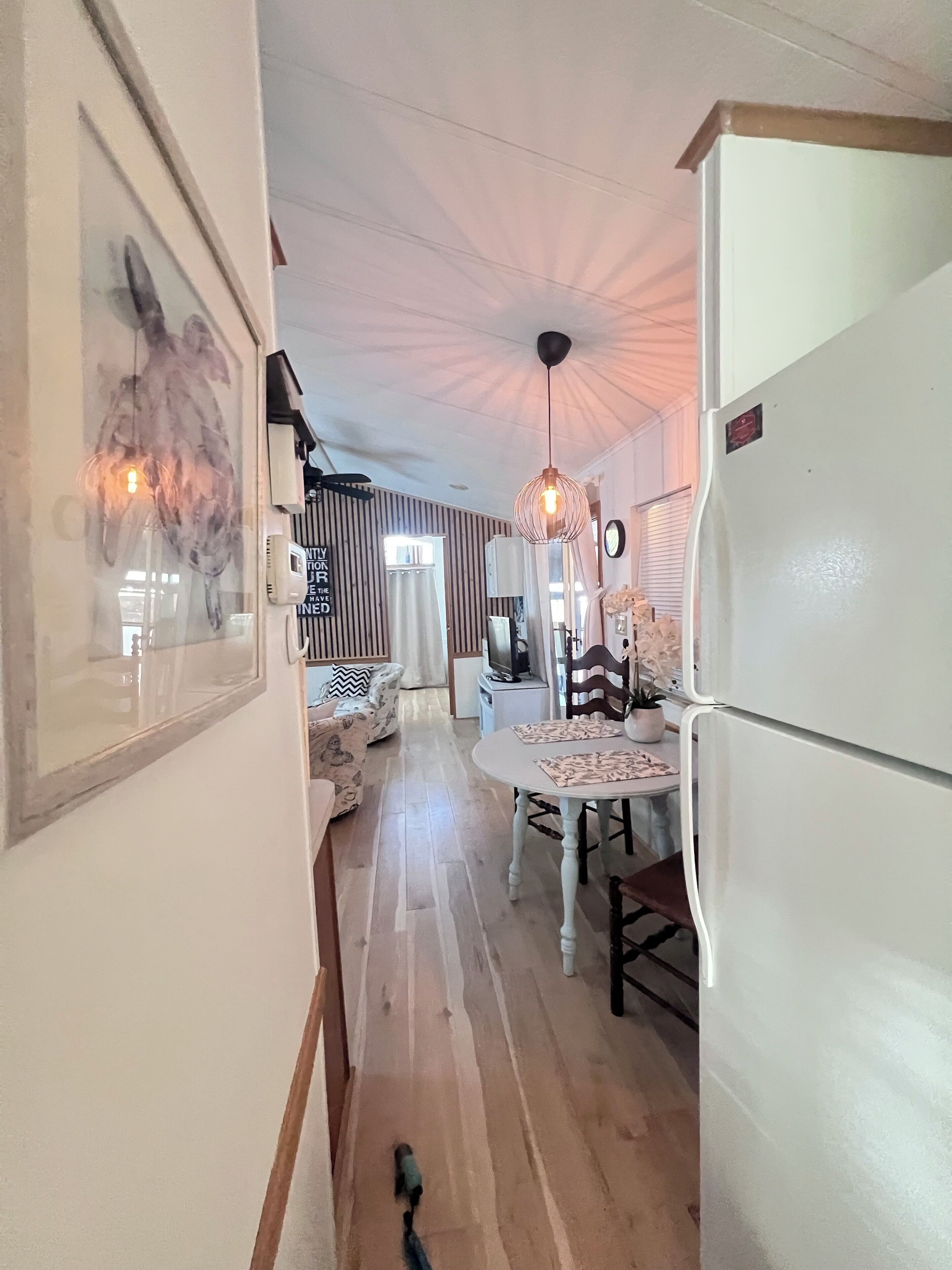 A narrow hallway with a dining area featuring a white table and chairs, a refrigerator, and a hanging light fixture.