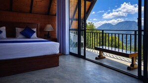 A bedroom with a large bed, a nightstand, a lamp, and a view of mountains through a balcony. - Forest Edge Hotel (Ginigathhena)