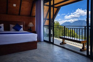 A bedroom with a large bed, a nightstand, a lamp, and a view of mountains through a balcony. - Forest Edge Hotel (Ginigathhena)