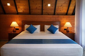 A hotel room with a wooden ceiling, brick walls, a large bed with white bedding and blue pillows, and two bedside lamps. - Forest Edge Hotel (Ginigathhena)