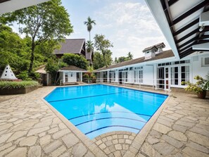 A swimming pool with stone tiles, surrounded by a white building with windows and a covered area. - Forest Edge Hotel (Ginigathhena)