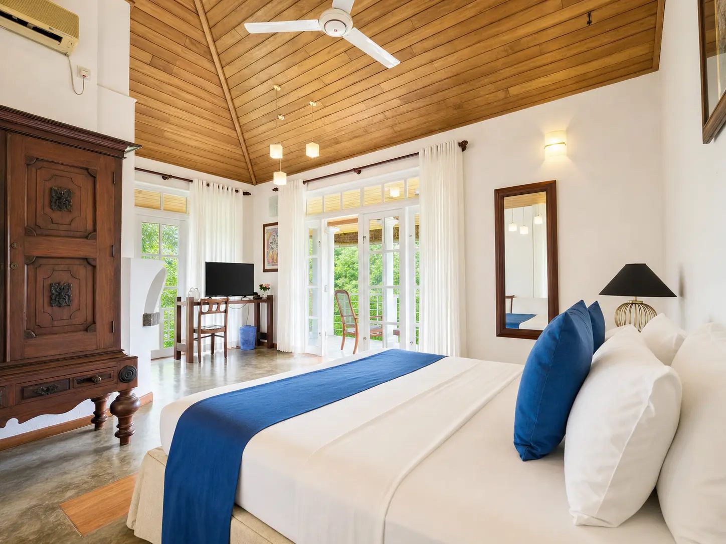 A hotel room with a wooden ceiling, a bed with blue and white bedding, a desk with a television, and a view of greenery outside.