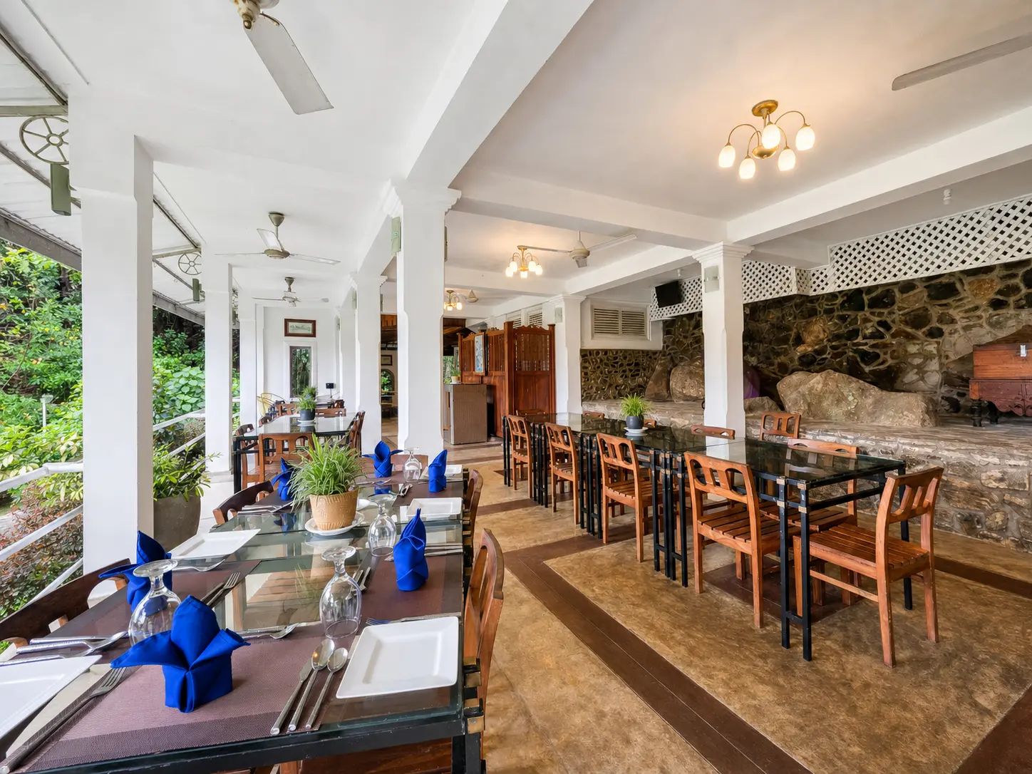 A spacious dining area with a glass table and wooden chairs, a stone wall, and a view of greenery outside.