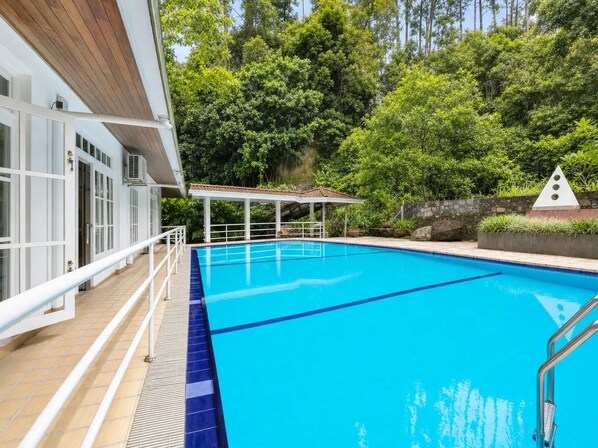 Una piscina azul cristalina con una cubierta de madera y un área de descanso techada, rodeada de abundante follaje.