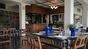 A restaurant with wooden furniture, a stone wall, and a kitchen area in the background. - Forest Edge Hotel (Ginigathhena)